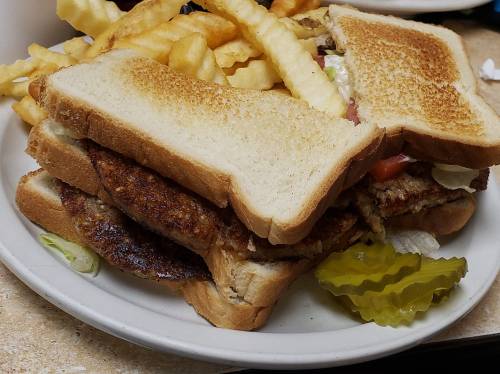 A plate with a stacked sandwich made of toasted bread, fried meat, lettuce, tomato, pickles, and a side of crinkle-cut fries.