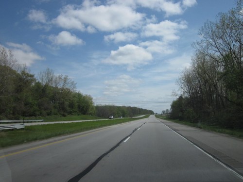 A wide, empty highway stretches into the distance under a partly cloudy sky, flanked by green trees.