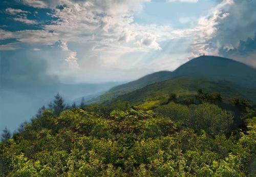 A scenic view of rolling green hills under a cloudy sky with rays of sunlight breaking through.