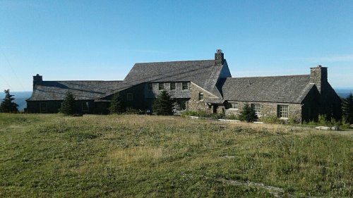A large stone house with a sloped roof, surrounded by grass and trees, set against a clear blue sky.