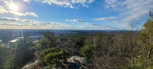 A panoramic view from a rocky hilltop, showcasing a landscape of trees, clouds, and distant valleys under a bright sun.