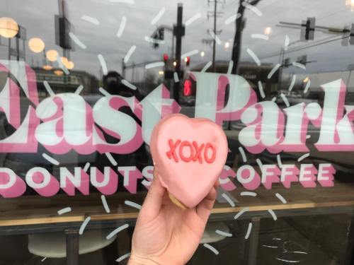A hand holds a heart-shaped pink donut with "XOXO" written on it, in front of a shop window that reads "East Park Donuts Coffee."