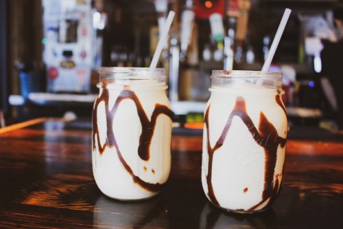 Two mason jars filled with creamy drinks, drizzled with chocolate syrup, sit on a wooden table.