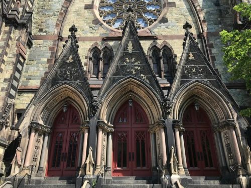 Gothic-style church entrance with three ornate arches and red doors, featuring intricate stonework and a large rose window above.