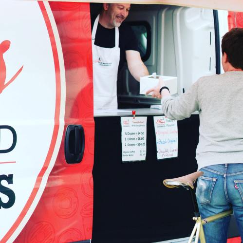 A vendor hands a customer a takeout box from a food truck, with menu prices visible on the side.
