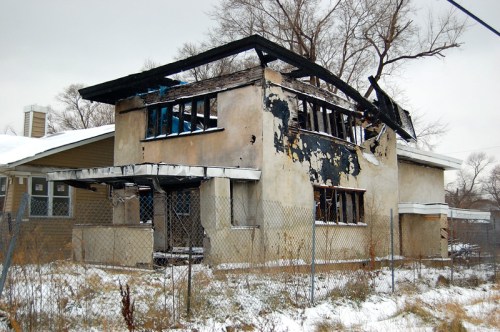 Abandoned, burned house with charred walls and a damaged roof, surrounded by overgrown grass and a chain-link fence.