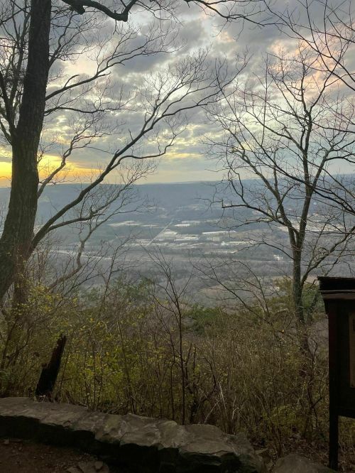 A scenic view from a mountain overlook, featuring trees, a cloudy sky, and a valley below at sunset.