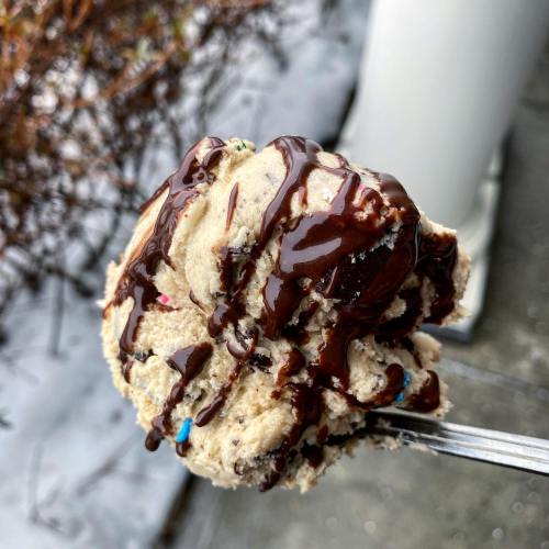 A close-up of a scoop of cookie dough ice cream drizzled with chocolate sauce, topped with colorful sprinkles.