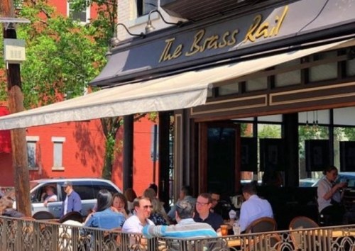 Outdoor dining scene at The Brass Rail, with patrons enjoying meals and drinks on a sunny day.