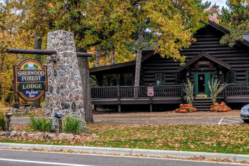 A rustic lodge surrounded by autumn foliage, featuring a stone sign and pumpkins in front.