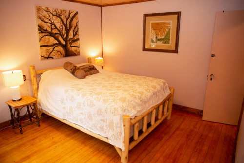 Cozy bedroom with a wooden bed, decorative pillows, lamps, and artwork on the walls. Warm wooden flooring.