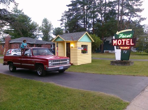 A red pickup truck parked near a small motel office with a bright "Vacancy" sign and trees in the background.