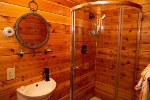 A wooden bathroom featuring a glass shower, round mirror, sink, and shelves with glasses. Warm, rustic decor.