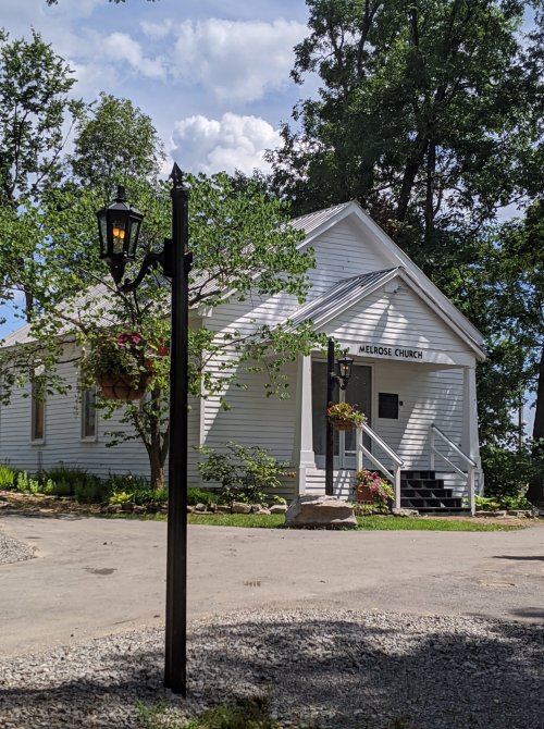 A small white church named Melrose Church surrounded by trees and flowers, with a lamp post in the foreground.