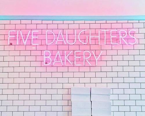 Neon sign reading "Five Daughters Bakery" on a white tiled wall, with stacked white boxes below.