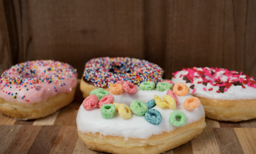 Four colorful donuts with various toppings, including sprinkles, cereal, and icing, arranged on a wooden surface.