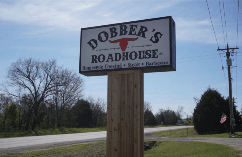 Sign for Dobber's Roadhouse, featuring "Homestyle Cooking, Steak, Barbecue" against a clear blue sky.