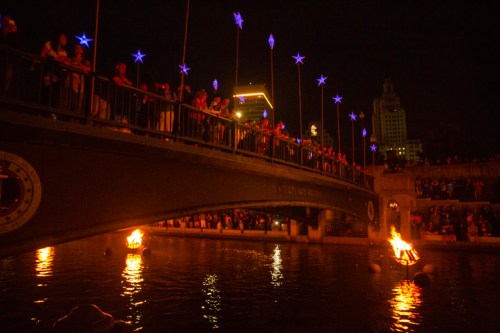 A crowd gathers on a bridge at night, with glowing stars and flames reflecting in the water below.