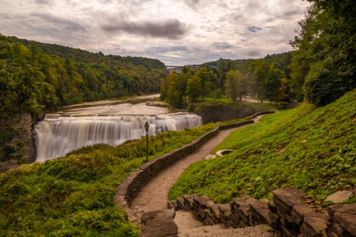 A scenic view of a waterfall surrounded by lush greenery and a winding path under a cloudy sky.