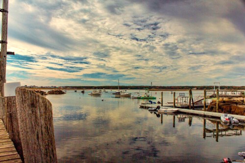 A serene harbor scene with boats docked, calm waters, and a cloudy sky reflecting over the landscape.