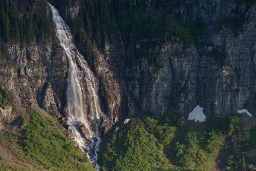 A majestic waterfall cascading down a rocky cliff, surrounded by lush greenery and patches of snow.