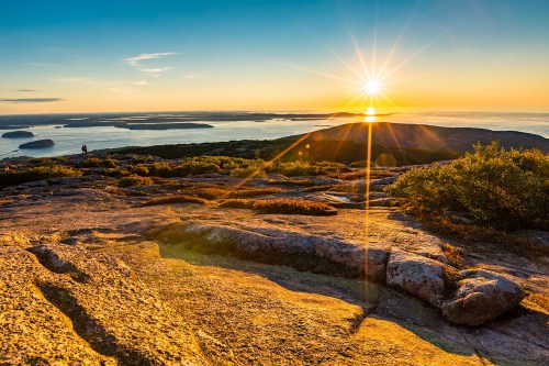 A stunning sunset over a rocky landscape, with a person silhouetted against the vibrant sky and distant islands.