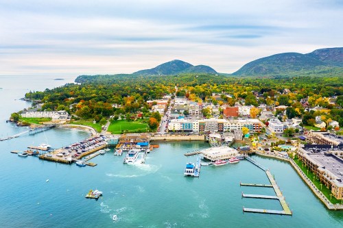 Aerial view of a coastal town with marinas, colorful autumn trees, and mountains in the background under a cloudy sky.