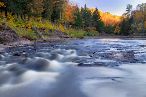 A serene river flows through a forest with vibrant autumn foliage, creating a peaceful natural scene.