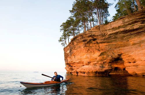 A person paddles a kayak near a rocky cliff with trees, reflecting the calm waters of a lake at sunset.
