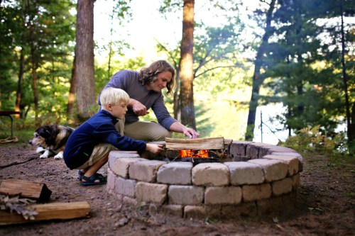A child and an adult add wood to a campfire in a forested area, with a dog resting nearby and a lake in the background.