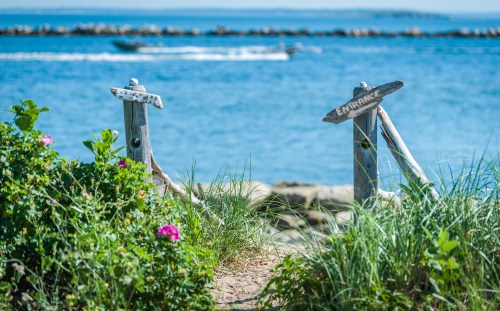 A pathway leads to the water, marked by signs reading "Beach" and "Entrance," surrounded by greenery and a boat in the distance.