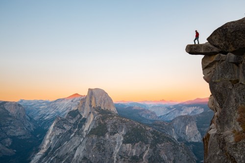 A lone hiker stands on a rocky ledge overlooking a vast mountain landscape at sunset.