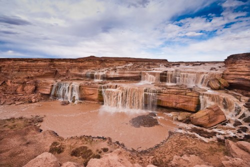 A scenic view of cascading waterfalls over rocky cliffs, surrounded by a cloudy sky and earthy terrain.