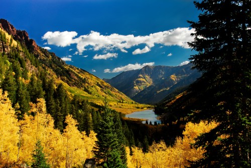 A scenic view of a valley with golden autumn trees, mountains, and a calm lake under a blue sky with fluffy clouds.