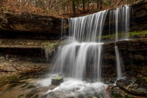 A serene waterfall cascades over rocky cliffs, surrounded by lush greenery and autumn leaves.