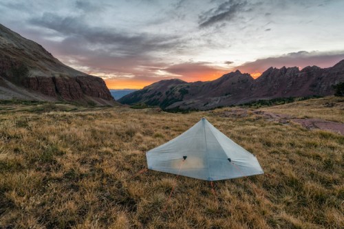A tent set up in a grassy valley, surrounded by mountains at sunset, with a colorful sky above.