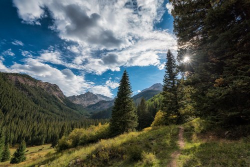 A scenic mountain landscape with tall trees, a winding path, and a bright sun peeking through clouds.