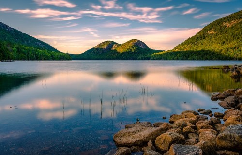 A serene lake reflects two mountains under a colorful sky, surrounded by lush greenery and rocky shores.
