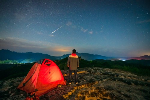 A person stands near a glowing tent, gazing at a starry sky with shooting stars over a mountainous landscape.