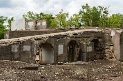 A weathered concrete structure with arches, surrounded by sparse vegetation and a cloudy sky.