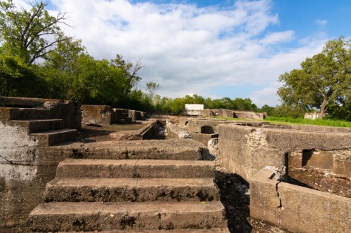 Ruins of stone structures with steps, surrounded by greenery and under a partly cloudy sky.