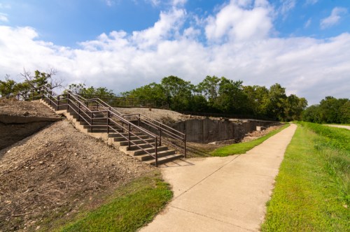 A concrete path alongside a grassy area, leading to a set of stairs descending into a partially eroded landscape.