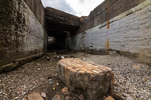 A desolate, abandoned concrete tunnel with debris and a stone block in the foreground, under a cloudy sky.