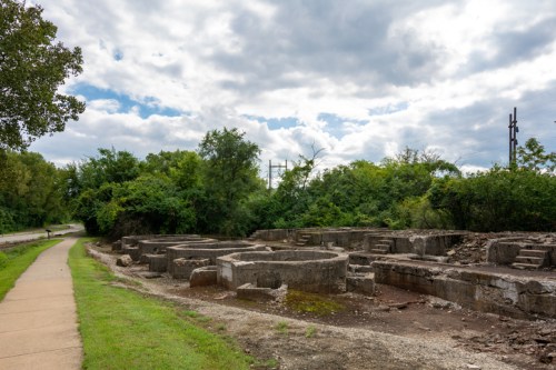 Ruins of concrete structures surrounded by greenery, with a pathway and cloudy sky in the background.