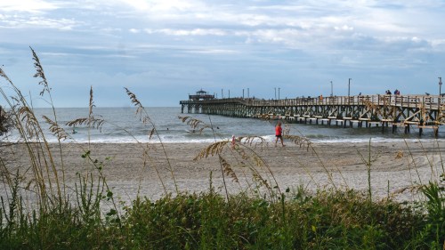 A sandy beach with tall grass in the foreground and a wooden pier extending into the calm sea under a cloudy sky.