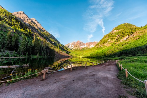 Scenic view of a mountain landscape with a clear lake, lush greenery, and a dirt path under a blue sky.