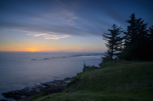 Sunset over a calm ocean, with silhouettes of trees and rocky shoreline in the foreground. Soft clouds in the sky.