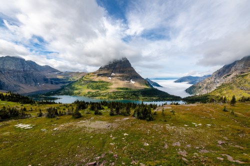 A scenic mountain landscape with lush greenery, a lake, and clouds partially covering the peaks under a blue sky.