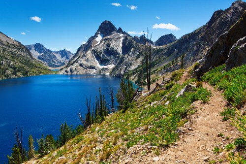 A scenic mountain landscape with a clear blue lake, rocky terrain, and lush greenery under a bright blue sky.