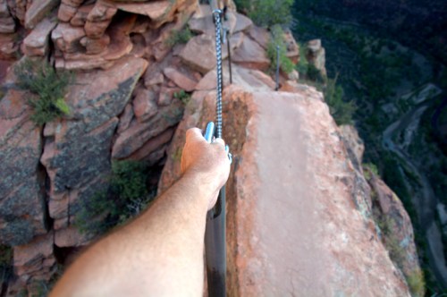 A hand grips a chain along a narrow rocky ledge overlooking a canyon.